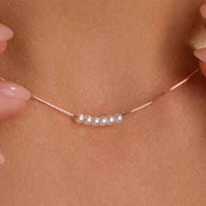 May include: A delicate rose gold necklace featuring a series of six small, white pearls. The necklace has a fine chain and is displayed against a blurred skin tone background. The pearls are evenly spaced along the chain.