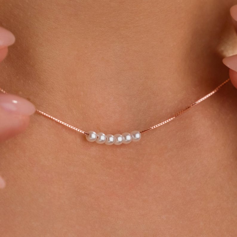 May include: A delicate rose gold necklace featuring a series of six small, white pearls. The necklace has a fine chain and is displayed against a blurred skin tone background. The pearls are evenly spaced along the chain.