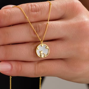 May include: Gold-colored necklace featuring a pendant with a white center and a cat silhouette. The pendant is surrounded by gold stars. The necklace is held in a hand, showcasing the delicate chain and pendant design.