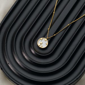 May include: A gold-colored necklace with a pendant featuring a cat silhouette and star cutouts on a white background. The necklace is displayed on a black, curved surface.