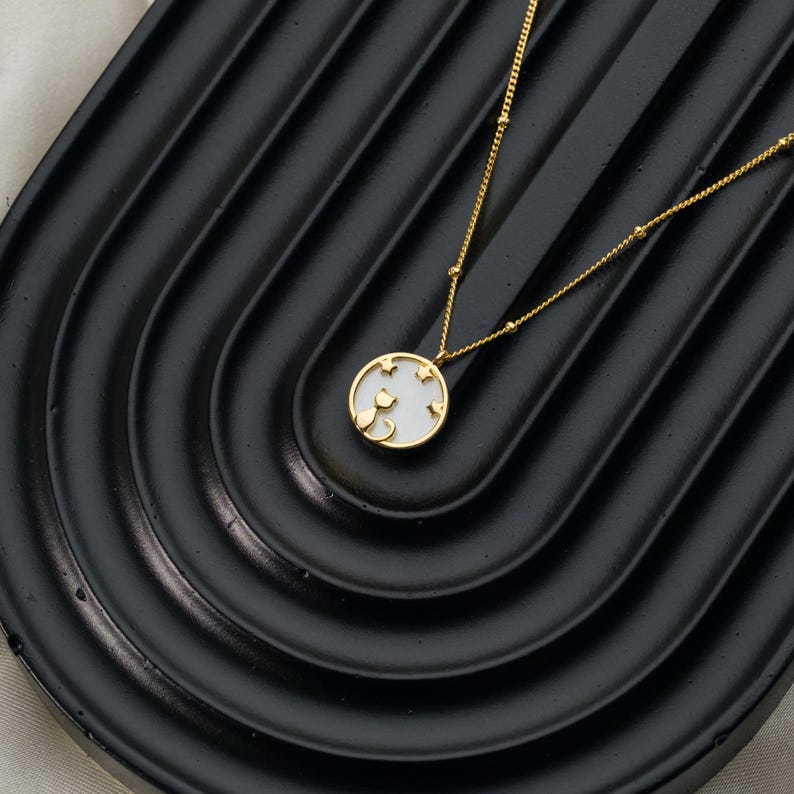 May include: A gold-colored necklace with a pendant featuring a cat silhouette and star cutouts on a white background. The necklace is displayed on a black, curved surface.