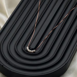 May include: A delicate necklace featuring a thin, rose-gold chain. The necklace has a series of small, white pearls clustered together. The jewelry is displayed on a black, textured surface, creating a contrast between the colors.