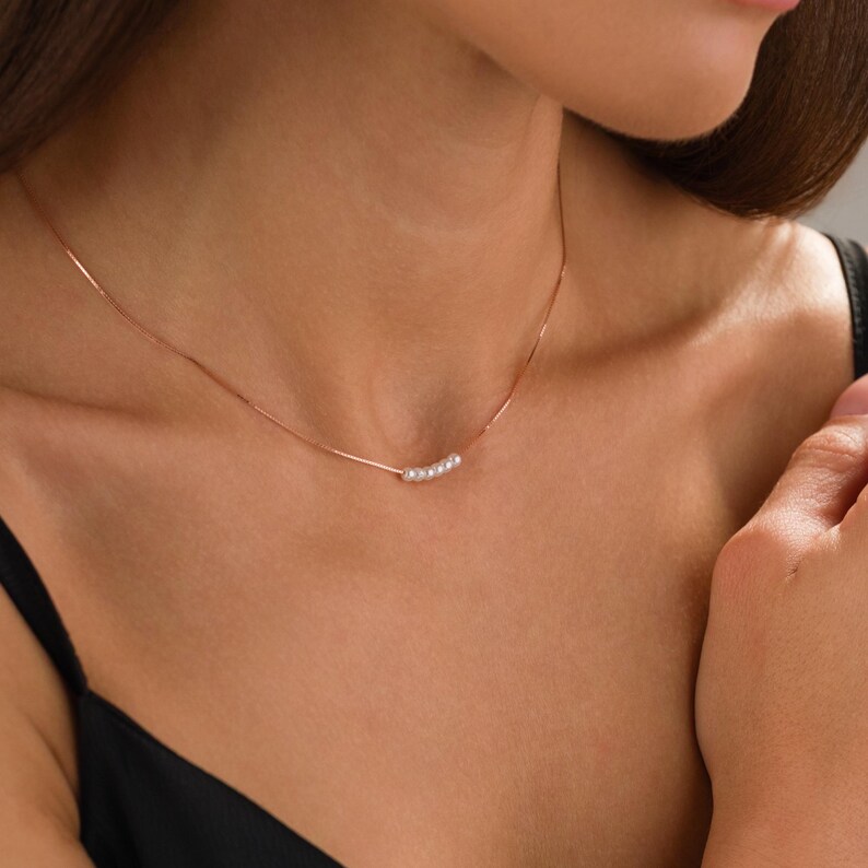 May include: A delicate necklace with a thin, rose-gold chain. The necklace features a series of small, white pearls arranged in a gentle curve. The jewelry is displayed against a dark background.