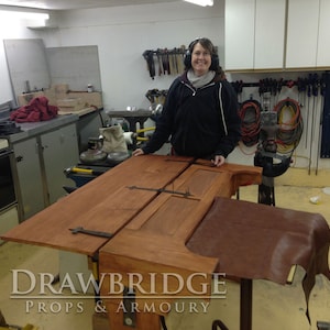Peut inclure: Une porte en bois avec des charni&egrave;res en m&eacute;tal et un morceau de cuir brun sur un &eacute;tabli dans un atelier. Le texte "Drawbridge Props & Armoury" est visible sur l'image.