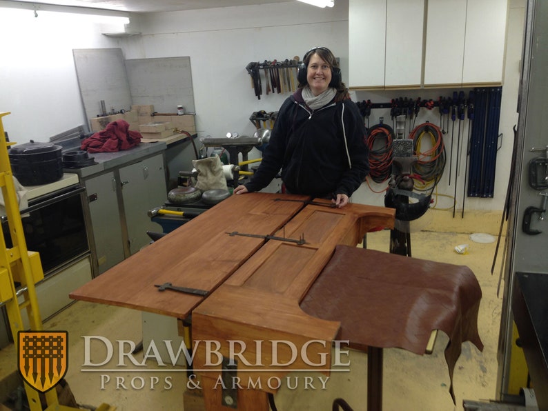 Peut inclure: Une porte en bois avec des charni&egrave;res en m&eacute;tal et un morceau de cuir brun sur un &eacute;tabli dans un atelier. Le texte "Drawbridge Props & Armoury" est visible sur l'image.