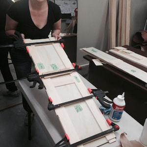 Peut inclure: Une femme utilise des serre-joints pour assembler un cadre de porte en bois. Le cadre est compos&eacute; de plusieurs morceaux de bois qui sont coll&eacute;s ensemble. La femme porte une chemise noire et un jean. Il y a un flacon de colle &agrave; bois sur la table.