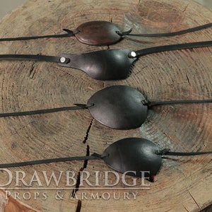 Four different black eye patches with leather thonging siting on a natural wooden tree stump from Drawbridge Props and Armoury.
