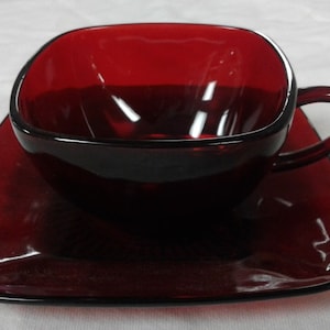 May include: A square red glass teacup and saucer set. The cup has a handle and the saucer is slightly angled.