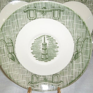 May include: A green and white ceramic saucer with a green design of a butter churn and wheelbarrows. The saucer has a raised rim and a smooth surface.