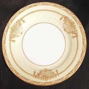 May include: A round, off-white plate with a gold rim and decorative gold filigree patterns. The plate features a central white circle and a gold border with a repeating leaf design. The plate's design is symmetrical and elegant.
