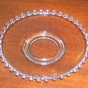 May include: A clear glass saucer with a beaded edge. The saucer has a smaller, raised center circle.
