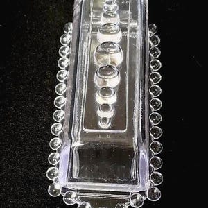 May include: A clear, rectangular glass butter dish with a series of graduated, spherical accents down the center. The edges are lined with small, clear, spherical beads. The dish is set against a black background.