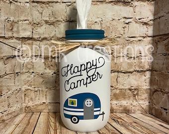 May include: White painted glass jar with a teal lid and a blue and white camper design. The jar has the text "Happy Camper" written on it. The jar is filled with tissues and has a brown twine bow around the lid.