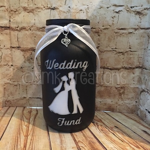 May include: Black glass jar with a white ribbon and a silver heart charm. The jar is decorated with a white silhouette of a bride and groom and the words "Wedding Fund".