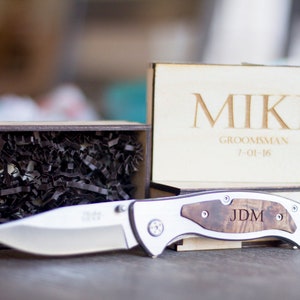 Może przedstawiać: Drewniane pudełko z personalizowanym grawerem z imieniem "Mike" na pokrywie. Pudełko zawiera srebrny scyzoryk z drewnianym uchwytem i wygrawerowanymi inicjałami "JDM". Pudełko jest prezentem dla świadka.