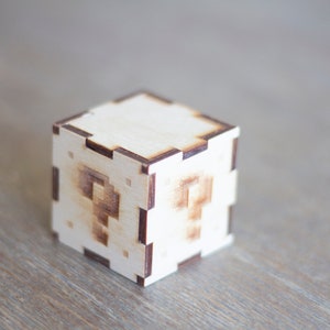 May include: A wooden box with a question mark design on each side. The box is made of light brown wood and has a dark brown border.