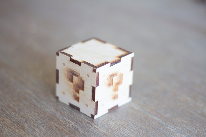 May include: A wooden box with a question mark design on each side. The box is made of light brown wood and has a dark brown border.