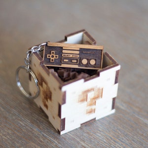 May include: A wooden keychain shaped like a vintage video game controller, with the words "Select Start" and "Nintendo" engraved on it. The keychain is in a small wooden box with a pixelated design.