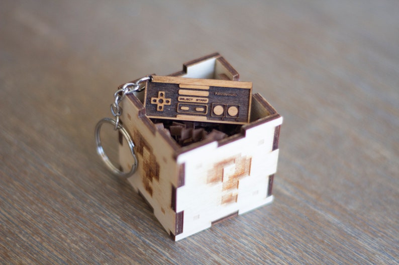 May include: A wooden keychain shaped like a vintage video game controller, with the words "Select Start" and "Nintendo" engraved on it. The keychain is in a small wooden box with a pixelated design.