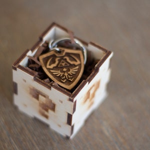 May include: A wooden box with a brown wood keychain inside. The keychain is shaped like a shield with a design of a bird and a triangle. The box is made of light wood and has a laser-cut design.