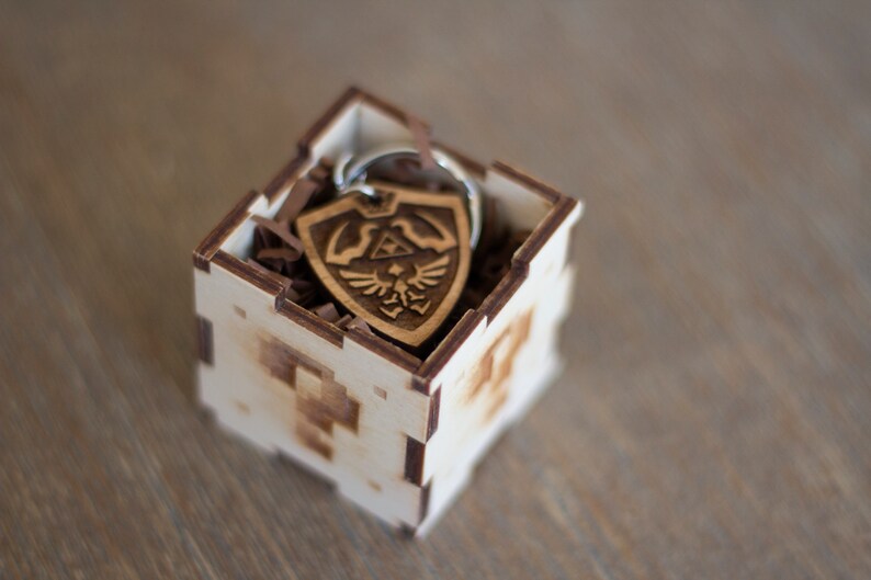 May include: A wooden box with a brown wood keychain inside. The keychain is shaped like a shield with a design of a bird and a triangle. The box is made of light wood and has a laser-cut design.