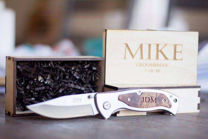 Może przedstawiać: Drewniane pudełko z wygrawerowanym imieniem "Mike" oraz tekstem "Groomsman 7-01-16". Pudełko zawiera srebrny scyzoryk z drewnianym uchwytem i wygrawerowanymi inicjałami "JDM".