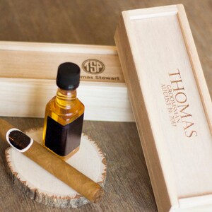 May include: A wooden box with the text "THOMAS GROOMSMAN AUGUST 19, 2017" engraved on the top. The box is closed and sitting on a wooden surface. A smaller wooden box with the initials "TSP" and the name "Thomas Stewart" engraved on the top is sitting next to the larger box. A small bottle of amber liquid with a black label is sitting on a wooden round slice. A brown cigar is sitting on the wooden round slice next to the bottle.