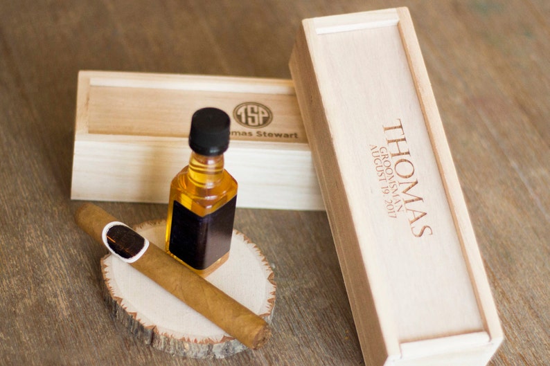 May include: A wooden box with the text "THOMAS GROOMSMAN AUGUST 19, 2017" engraved on the top. The box is closed and sitting on a wooden surface. A smaller wooden box with the initials "TSP" and the name "Thomas Stewart" engraved on the top is sitting next to the larger box. A small bottle of amber liquid with a black label is sitting on a wooden round slice. A brown cigar is sitting on the wooden round slice next to the bottle.