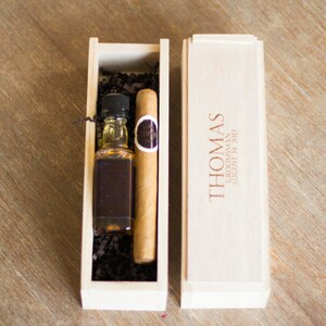 May include: A wooden box with a personalized inscription for "Thomas" and a date. The box contains a small bottle of liquor and a cigar.