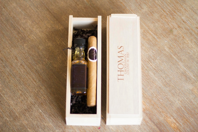 May include: A wooden box with a personalized inscription for "Thomas" and a date. The box contains a small bottle of liquor and a cigar.