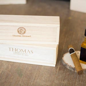 May include: Two wooden boxes with engraved text. The top box says "Thomas Stewart" with a monogram. The bottom box says "Thomas, Groomman, August 19, 2017". A small bottle of liquor and a cigar are on a wooden slice.
