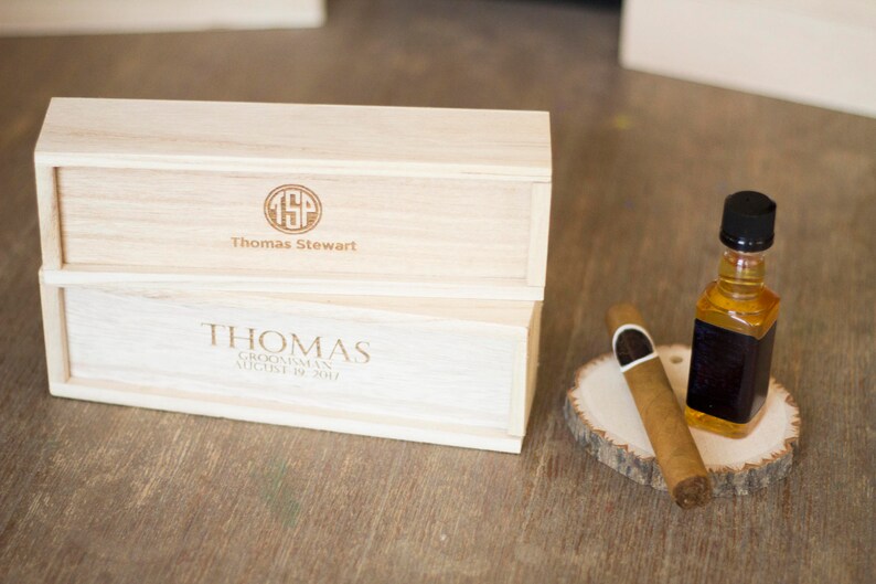 May include: Two wooden boxes with engraved text. The top box says "Thomas Stewart" with a monogram. The bottom box says "Thomas, Groomman, August 19, 2017". A small bottle of liquor and a cigar are on a wooden slice.