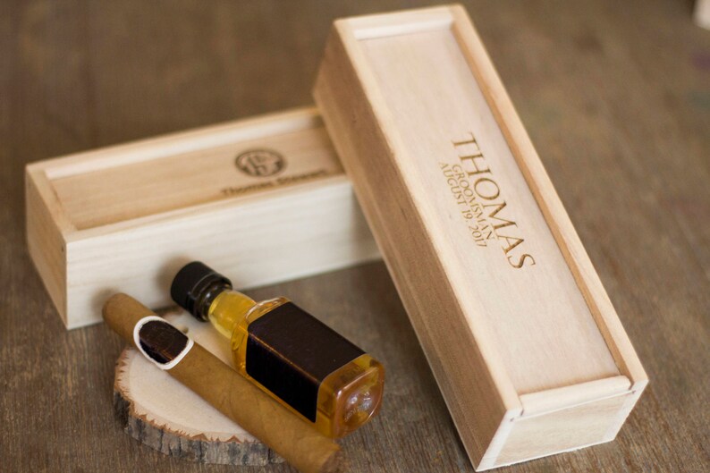 May include: Two wooden boxes with engraved text. The larger box has the text "THOMAS GROOMSMAN AUGUST 19, 2017". The smaller box has a cigar and a small bottle of liquor inside.