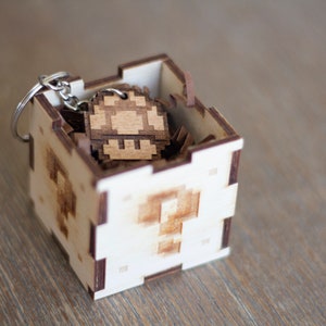 May include: A wooden box with a question mark design on the front, containing a wooden mushroom keychain. The keychain is made of light brown wood and has a pixelated design.