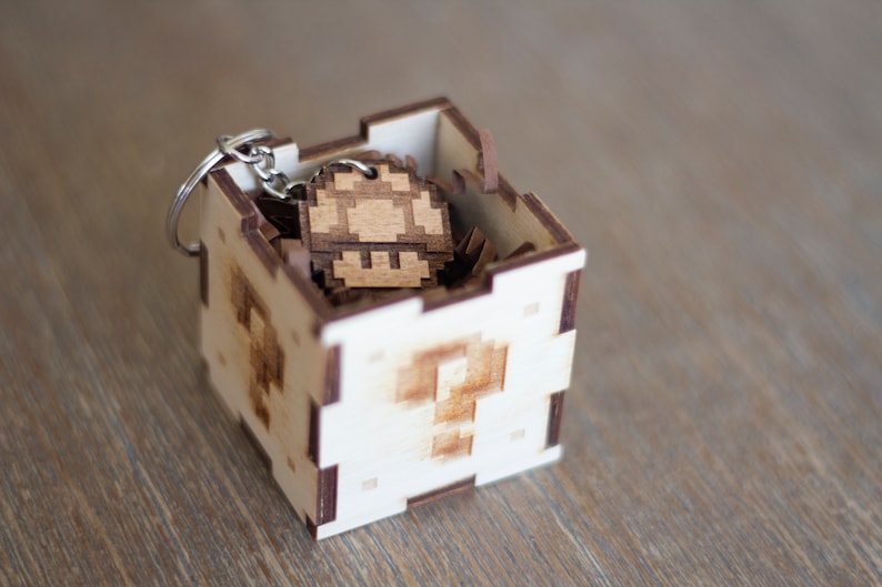 May include: A wooden box with a question mark design on the front, containing a wooden mushroom keychain. The keychain is made of light brown wood and has a pixelated design.