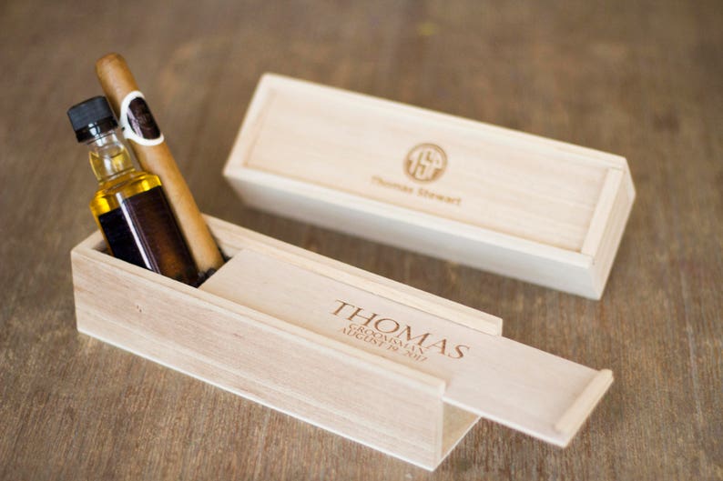 May include: A wooden box with a sliding lid, engraved with the name "Thomas" and the text "Groom's Man August 10, 2019". The box contains a small bottle of liquor and a cigar.