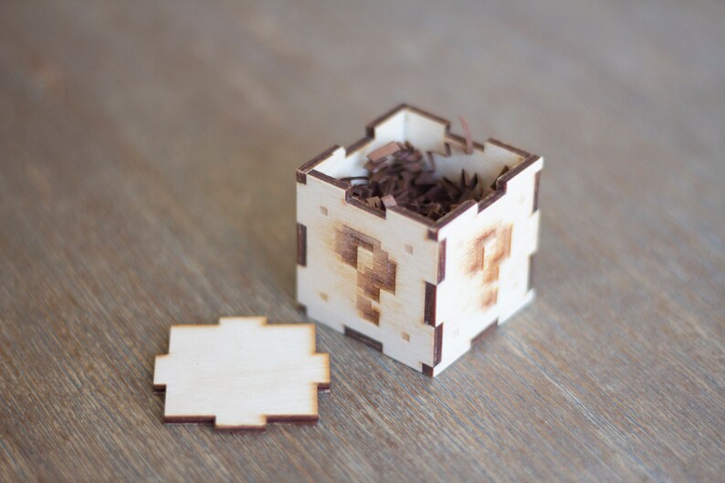 May include: A small wooden box with a question mark design on each side. The box is open and filled with brown shredded paper.