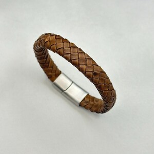 May include: A brown leather bracelet with a woven design. The bracelet features a silver-coloured magnetic clasp. The leather is a medium brown and has a worn appearance.