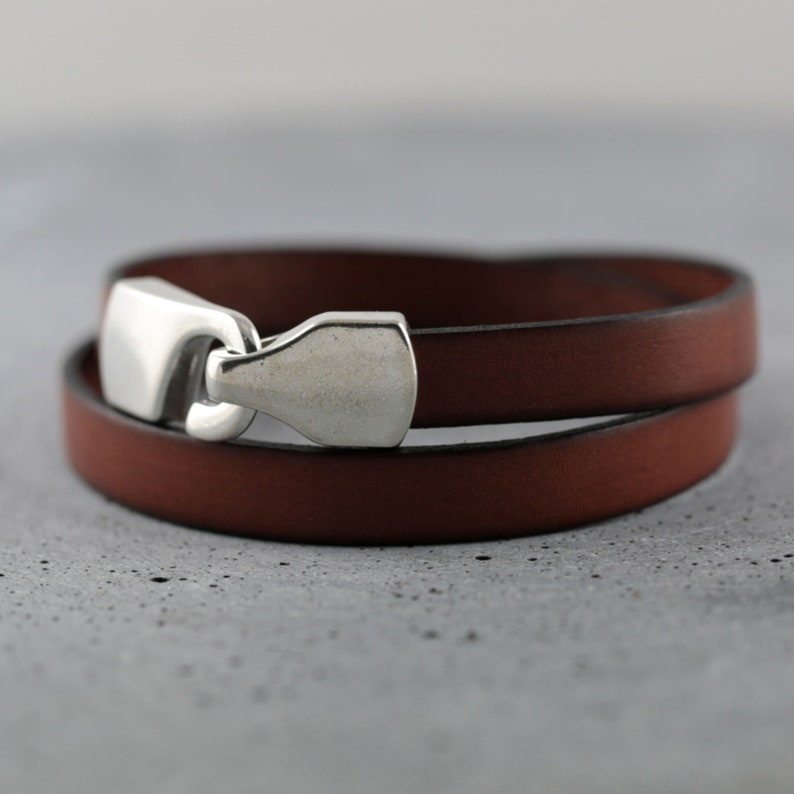 May include: A brown leather wrap bracelet with a silver-colored clasp. The bracelet is made of two layers of leather and has a simple, elegant design. The clasp is a rectangular shape with rounded edges.