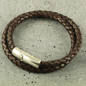 Puede incluir: Un brazalete de cuero marr&oacute;n con un dise&ntilde;o trenzado y un cierre plateado.