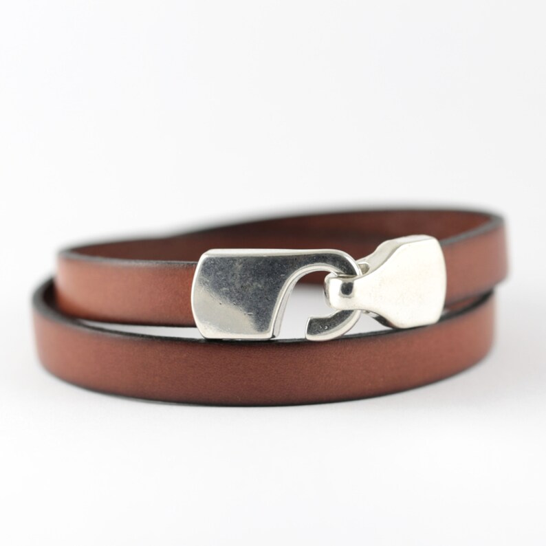 May include: A brown leather wrap bracelet with a silver-colored clasp. The bracelet is wrapped twice around, showcasing the rich color of the leather. The clasp is a simple, yet elegant design.