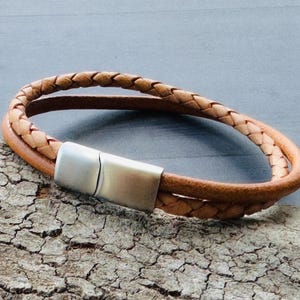 May include: A light brown leather bracelet with a braided top layer and a smooth, double-layered band. The bracelet has a silver-coloured magnetic clasp. The accessory is displayed on a textured wooden surface.