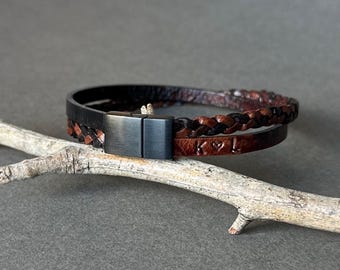 May include: A leather bracelet with a black metal clasp. The bracelet features a braided brown leather band and a smooth, dark brown leather band with the initials "K" and "L" and a heart symbol. The bracelet is displayed on a gray surface.