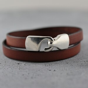 May include: A brown leather wrap bracelet with a silver-colored clasp. The bracelet is made of two bands of leather that wrap around the wrist. The clasp is a hook and eye style.