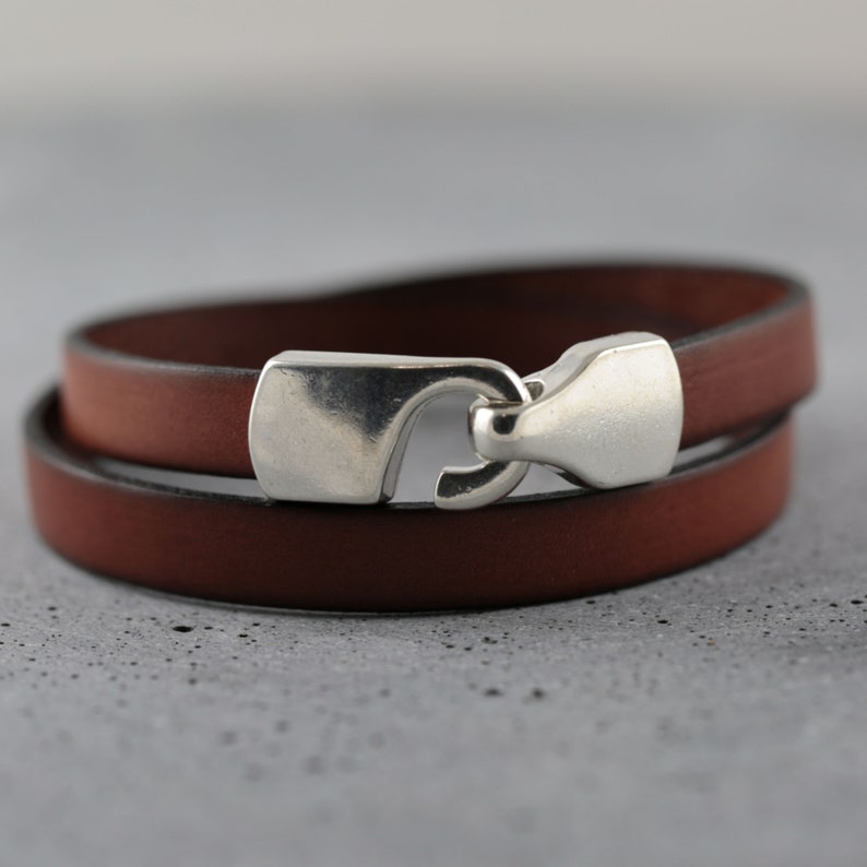 May include: A brown leather wrap bracelet with a silver-colored clasp. The bracelet is made of two bands of leather that wrap around the wrist. The clasp is a hook and eye style.
