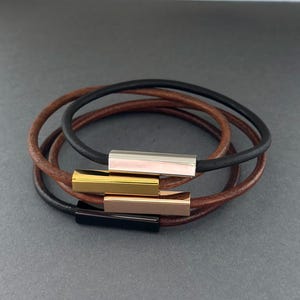May include: Four leather bracelets in a stack. The bracelets are in shades of brown and black. Each bracelet features a rectangular metal clasp in gold, silver, rose gold, and black.