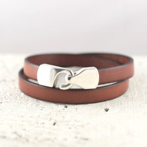 May include: A brown leather wrap bracelet with a silver-colored clasp. The bracelet is wrapped twice around and has a simple, minimalist design. The clasp is a hook and eye style.