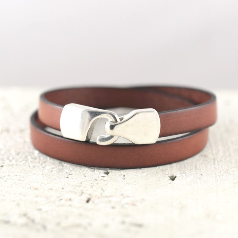 May include: A brown leather wrap bracelet with a silver-colored clasp. The bracelet is wrapped twice around and has a simple, minimalist design. The clasp is a hook and eye style.