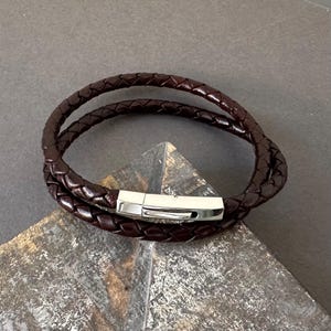 Puede incluir: Una pulsera de cuero trenzado marrón oscuro con un cierre plateado. La pulsera se envuelve dos veces. El cuero tiene un acabado pulido y el cierre es rectangular. Adecuado para una circunferencia de muñeca de aproximadamente 18 cm.