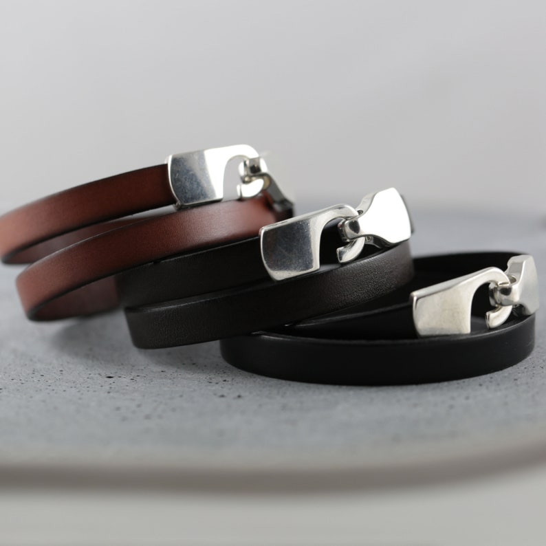 May include: Three leather bracelets in shades of brown and black, each with a silver-colored clasp. The bracelets are stacked on top of each other, showcasing the different colors and the design of the clasp.